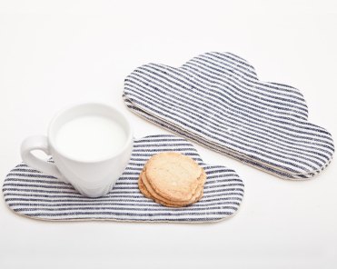 Still life photo of 2 striped coasters with a cloud shape, and a white cup with milk and biscuits on a side.